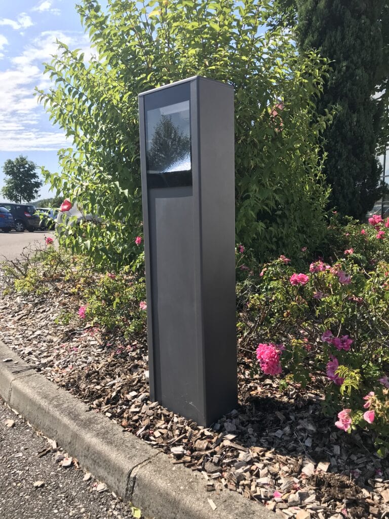 A gray, rectangular Lumea outdoor kiosk stands on mulch next to a curb, surrounded by flowering bushes and greenery under a partly cloudy sky.