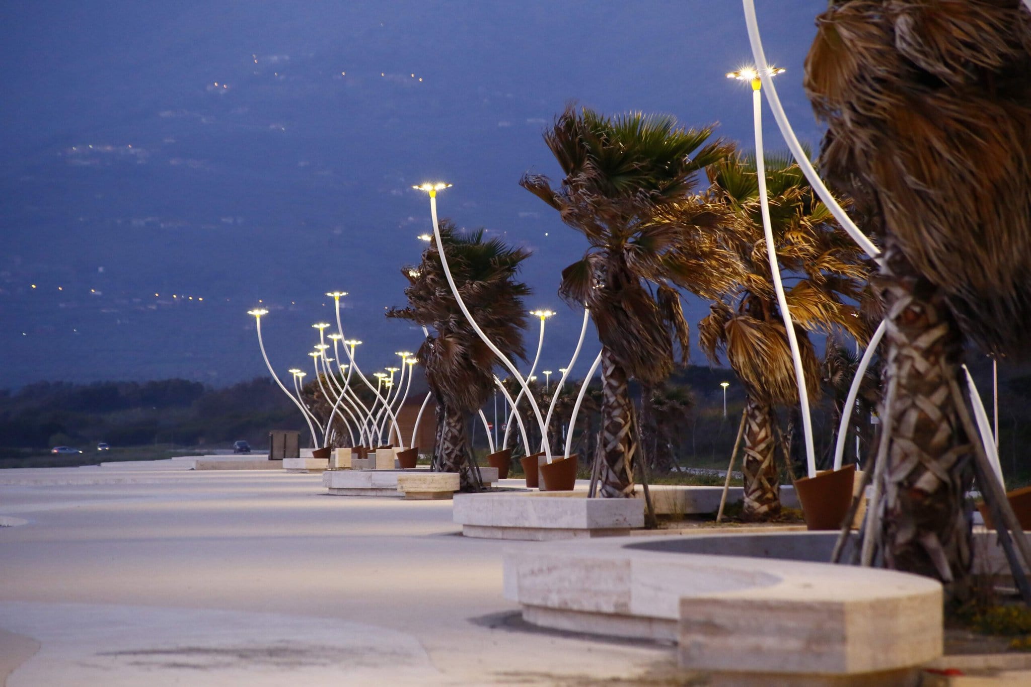 Curved streetlights line Margo’s coastal promenade at dusk, with palm trees bending in the wind and mountains faintly visible in the background.