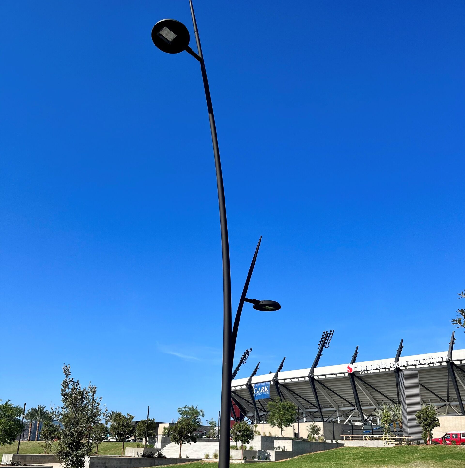 A modern black streetlight with two lamps on a sidewalk near a grassy area and stadium under a clear blue sky.