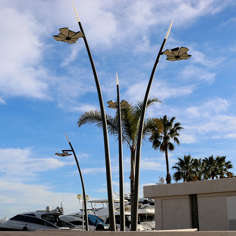 Tall, modern streetlights with flower-shaped fixtures stand near palm trees and docked boats under a partly cloudy sky.