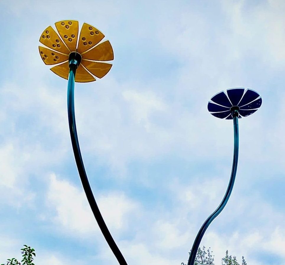 Two tall, curved metal sculptures by Margo, with flower-shaped tops—one yellow and one blue—stand against a cloudy sky.