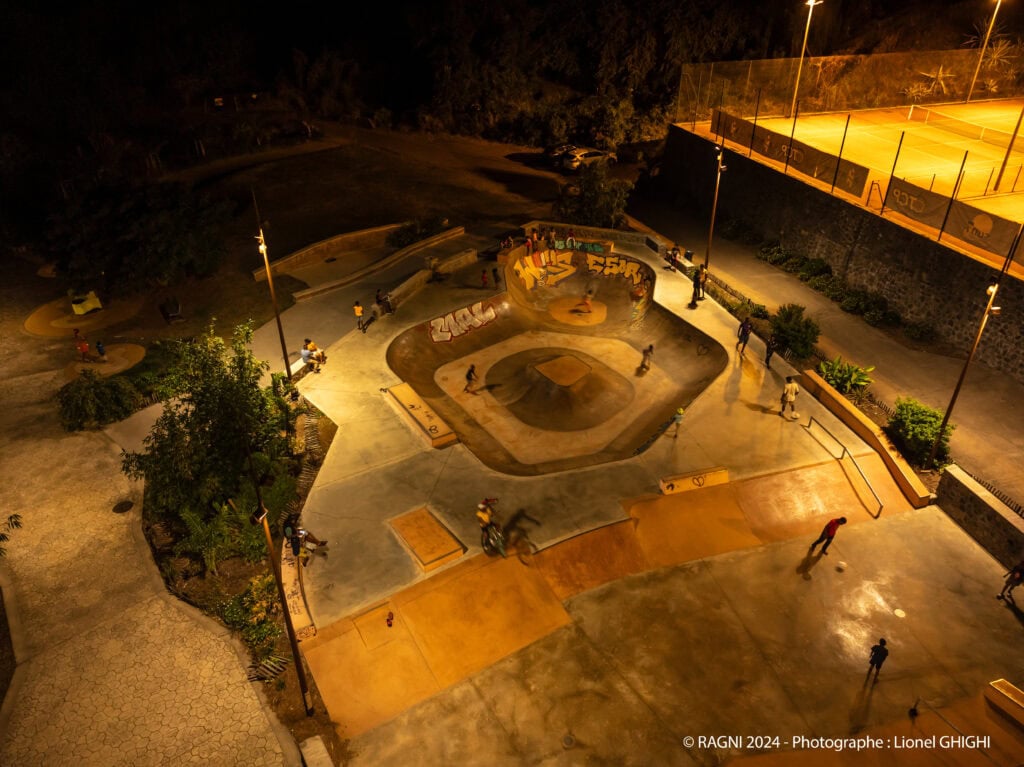 A well-lit skatepark at night with several people skating and walking; a brightly lit tennis court is visible at the top right, adding to the vibrant irys of evening activity.