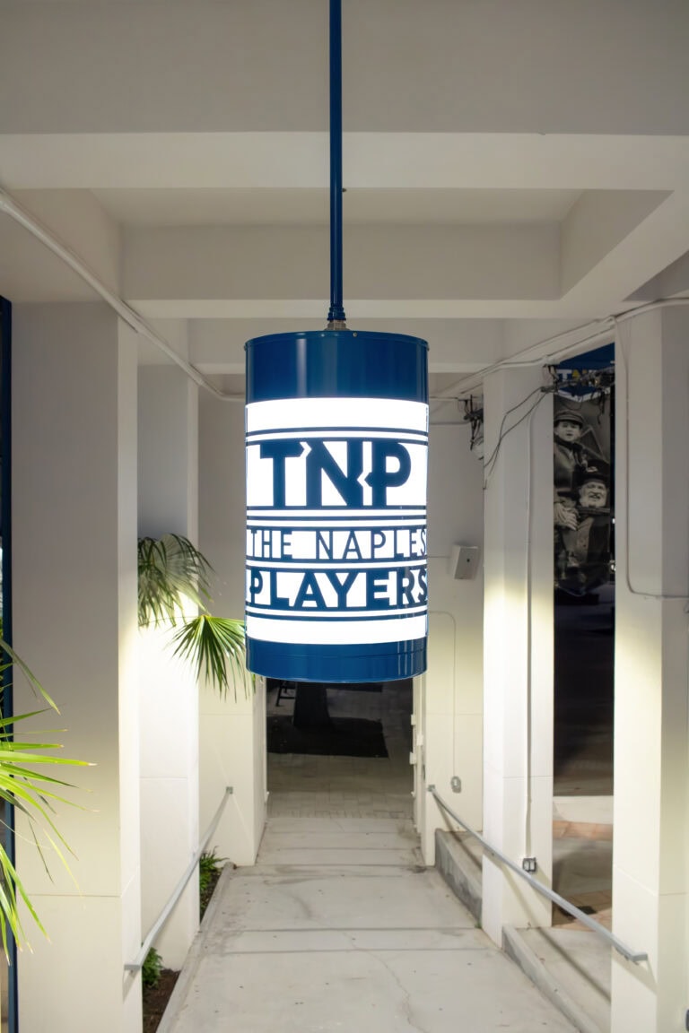 A cylindrical blue and white lampion-style hanging sign reads "TNP THE NAPLES PLAYERS" in a covered walkway, with plants and a mural visible nearby.