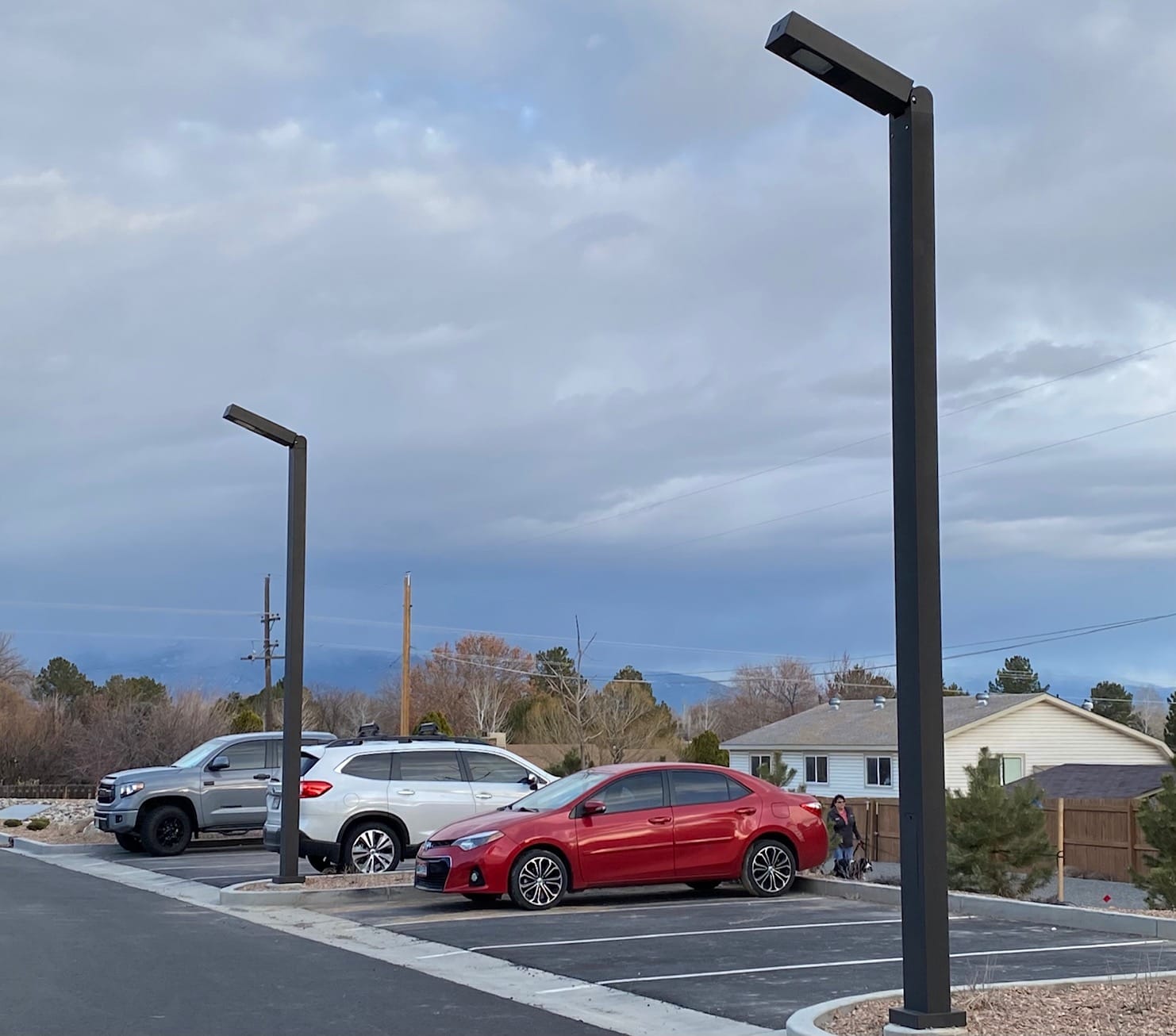 Three cars are parked outside under modern streetlights near residential homes, with a cloudy sky and trees in the background.
