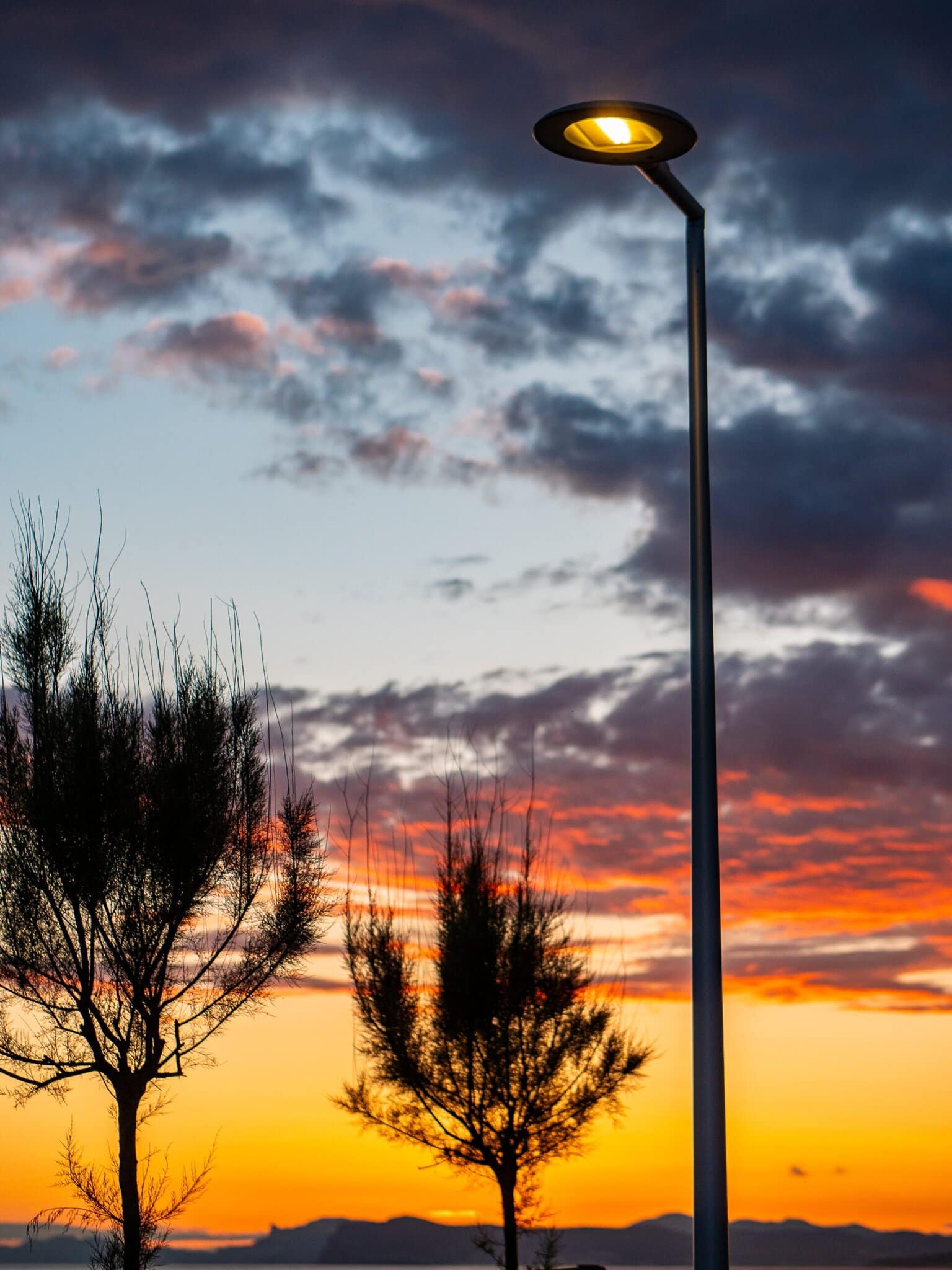 A streetlamp is lit at dusk, casting a gentle glow as silhouetted trees stand aro the base, with a vibrant sunset sky featuring clouds in orange, yellow, and purple hues in the background.