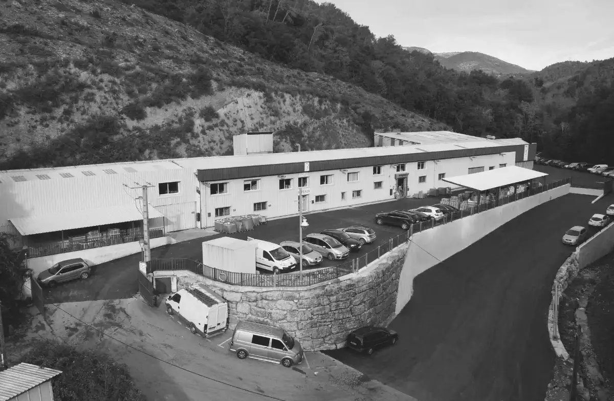 A large, rectangular industrial building with multiple parked cars in front, situated on a hillside with a paved road and surrounding trees.