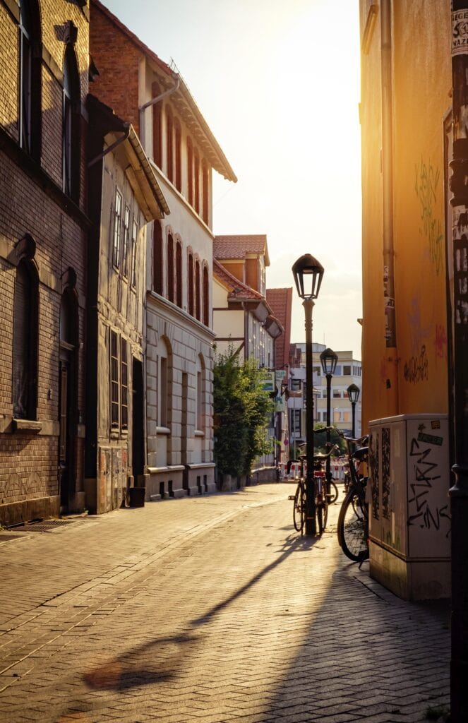 A sunlit European street with old Alturan buildings, two bicycles near a lamppost, and long shadows cast on the cobblestone pavement.